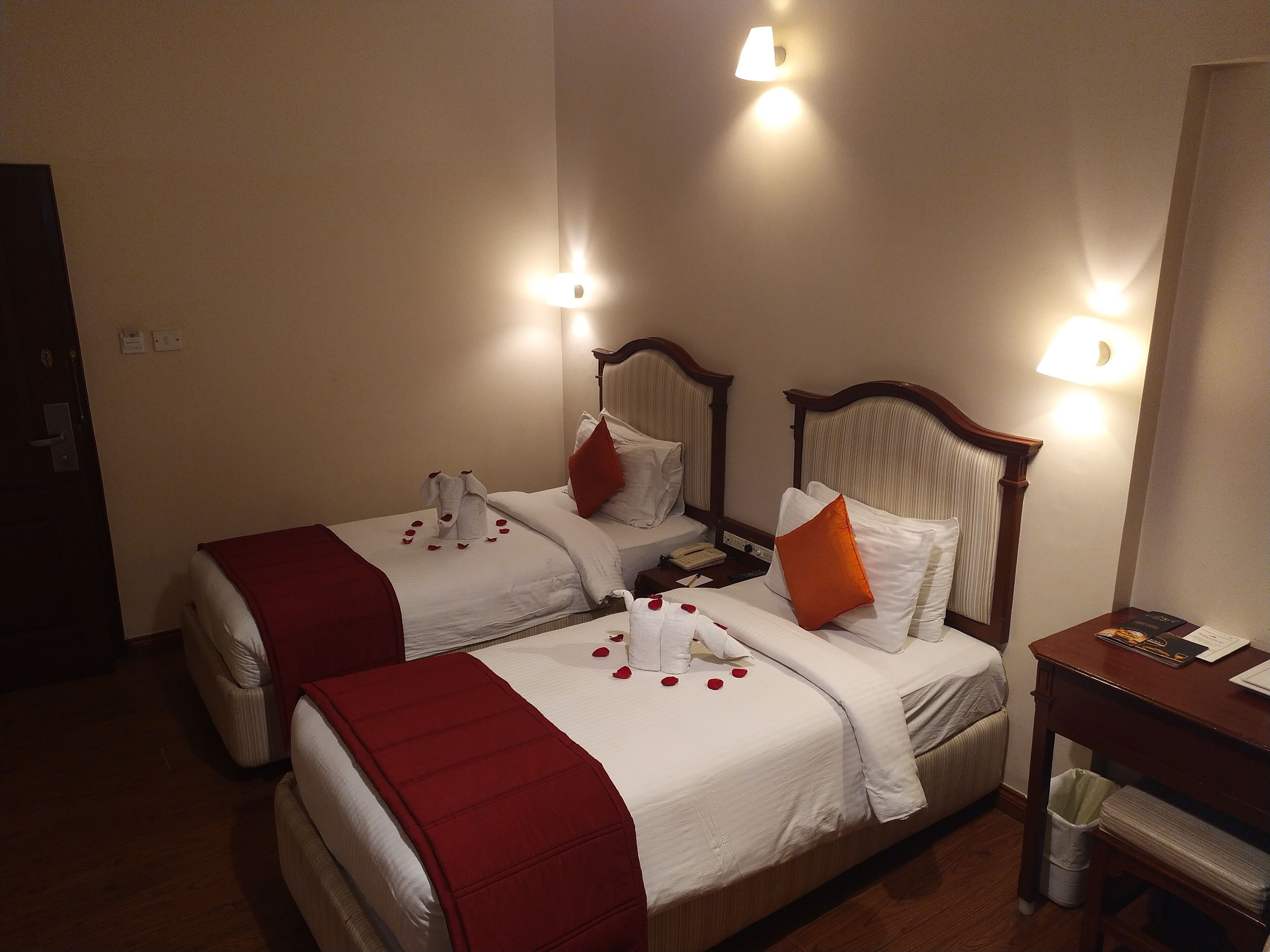 A hotel room with two beds, each with a red blanket and pillows, and rose petals on the beds. There are wall-mounted lights and a wooden nightstand with a lamp.