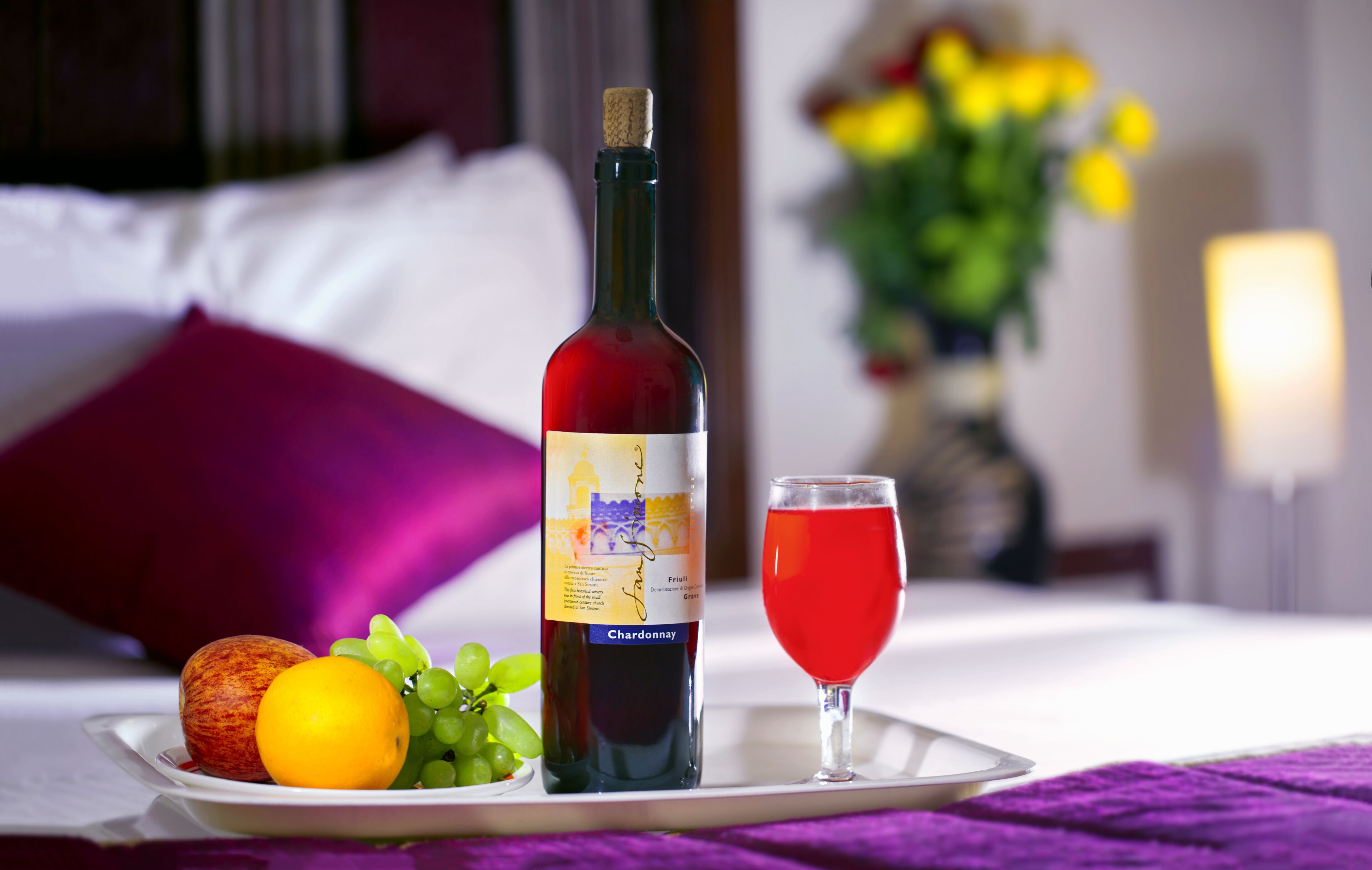 A bottle of Chardonnay wine, a glass of red wine, an apple, a lemon, and grapes on a tray on a bed with a purple pillow and a vase of yellow flowers in the background.