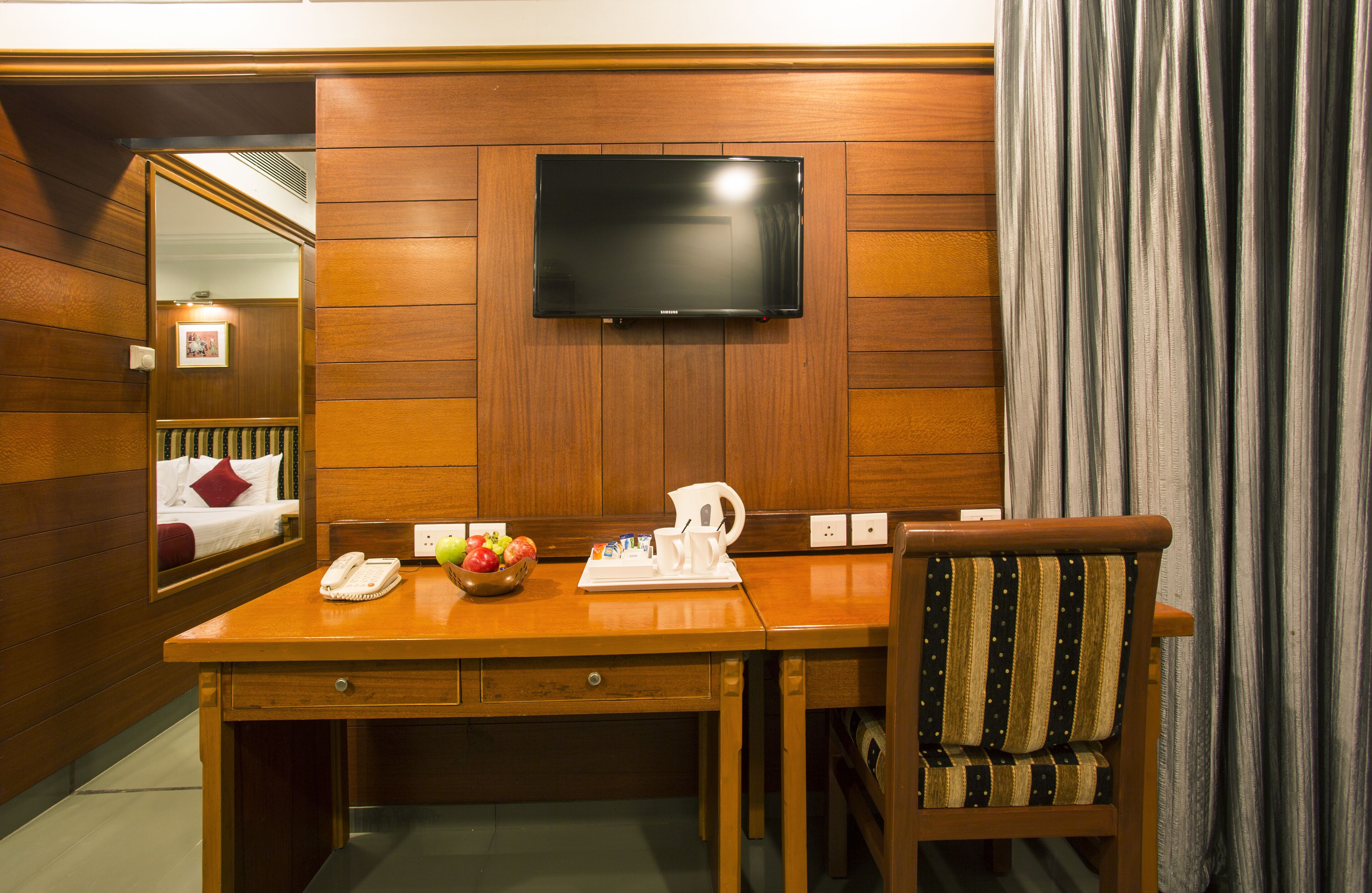 A hotel room with a wooden desk, a television mounted on the wall, a chair, a bedside table with a phone and a fruit bowl, and a mirror reflecting a bed with a pillow.