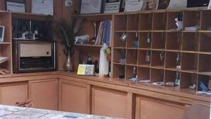 Una habitación con un escritorio de madera, estanterías con diversos objetos e iluminación fluorescente en el techo.