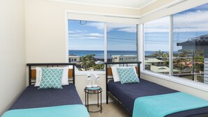 A room with two beds, a small table with lamps, and large windows offering a view of the ocean and buildings. - Beach Cottage (Forster)