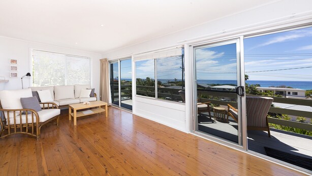 A spacious room with a wooden floor, a sofa, a table, and large sliding glass doors offering a view of the ocean and surrounding buildings. - Beach Cottage (Forster)