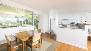 A modern kitchen with white cabinets, a central island, and a dining area with a wooden table and chairs. - Beach Cottage (Forster)