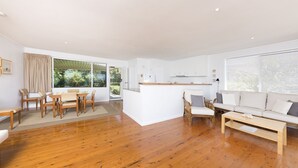 A spacious living area with a wooden floor, a dining set, a sofa, and a kitchen area with white cabinets. - Beach Cottage (Forster)