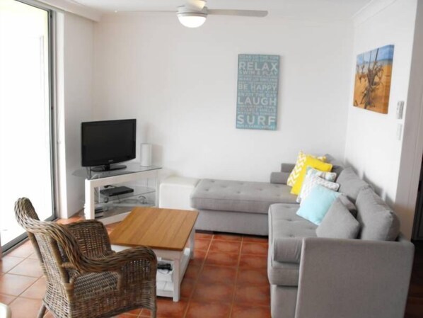 A living room with a grey sofa, a wicker chair, a coffee table, a TV on a stand, and a wall with motivational words.