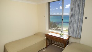 Une chambre avec deux lits simples, un bureau en bois agrémenté d’une plante et une grande fenêtre donnant sur la mer.