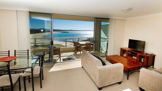 Ein Balkon mit Glastür und Blick auf den Strand, einem Fernsehtisch und einer Sitzecke mit Sofa und Sesseln.