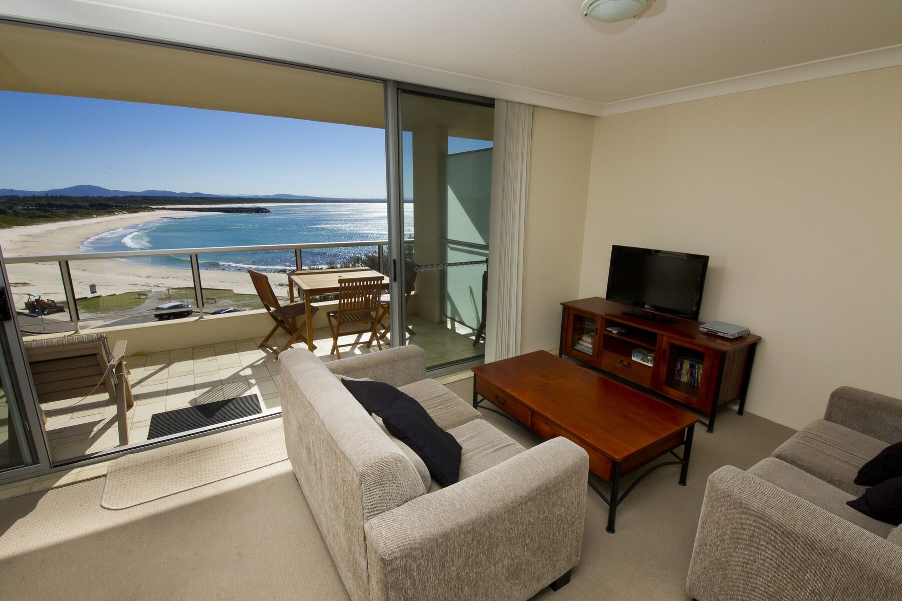 A balcony with a view of the beach and ocean. There is a sofa, a TV, and a wooden cabinet.