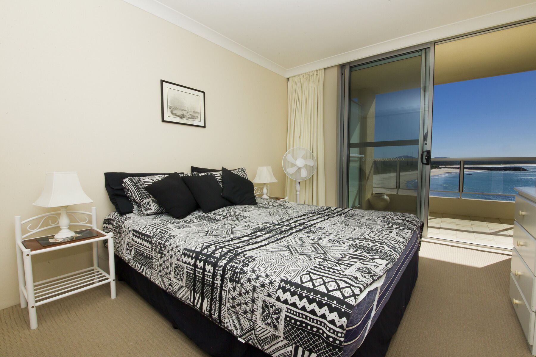 A hotel room with a large bed, a bedside table with a lamp, a fan, and a view of the ocean through a sliding glass door.