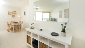 A bright, modern living room with a white console table, a large mirror, and a dining area with wooden chairs and a small table. - Beachpoint Unit 501 (Forster)