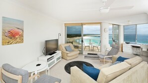 A modern living room with a TV, sofa, and chairs, a balcony with a view of the ocean, and a ceiling fan. - Beachpoint Unit 501 (Forster)