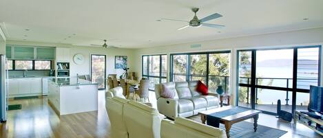 Ampla sala de estar com sofá, mesa de centro e televisão. Há uma cozinha com armários brancos e micro-ondas. Janelas grandes oferecem vista para o exterior.