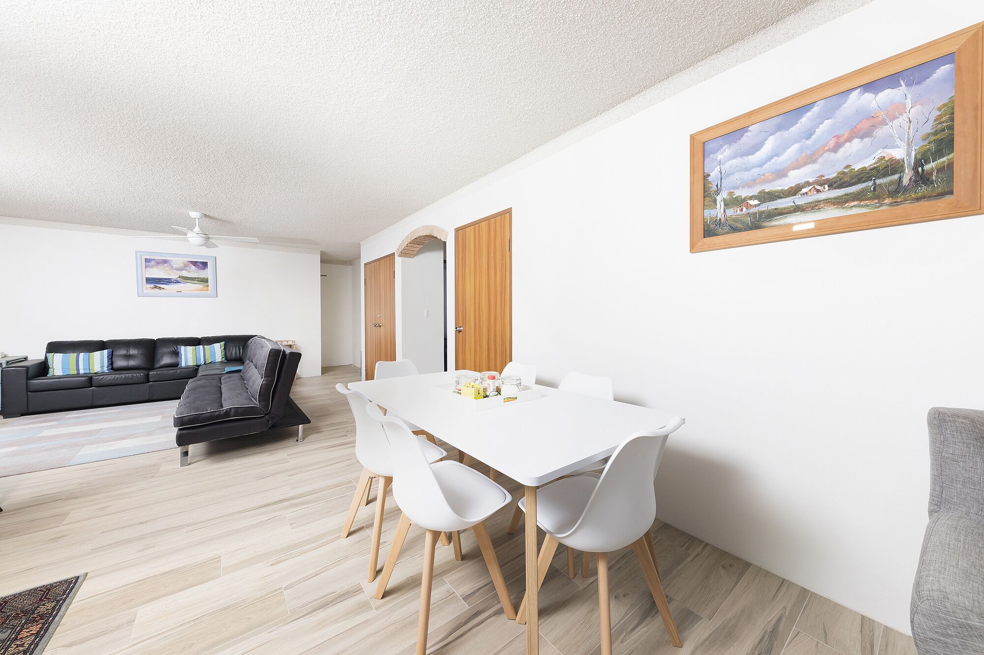 A modern living room with a black leather sofa, a white dining table with four chairs, and a painting on the wall.