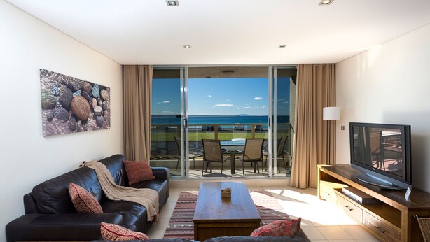 A modern living room with a leather sofa, a coffee table, and a TV on a wooden stand. A large window offers a view of the sea and a balcony with outdoor furniture. - Ebbtide Unit 04 (Forster)