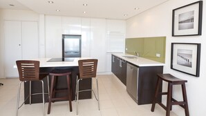 A modern kitchen with white cabinets, a stainless steel refrigerator, a large island with a sink, and two bar stools. - Ebbtide Unit 04 (Forster)