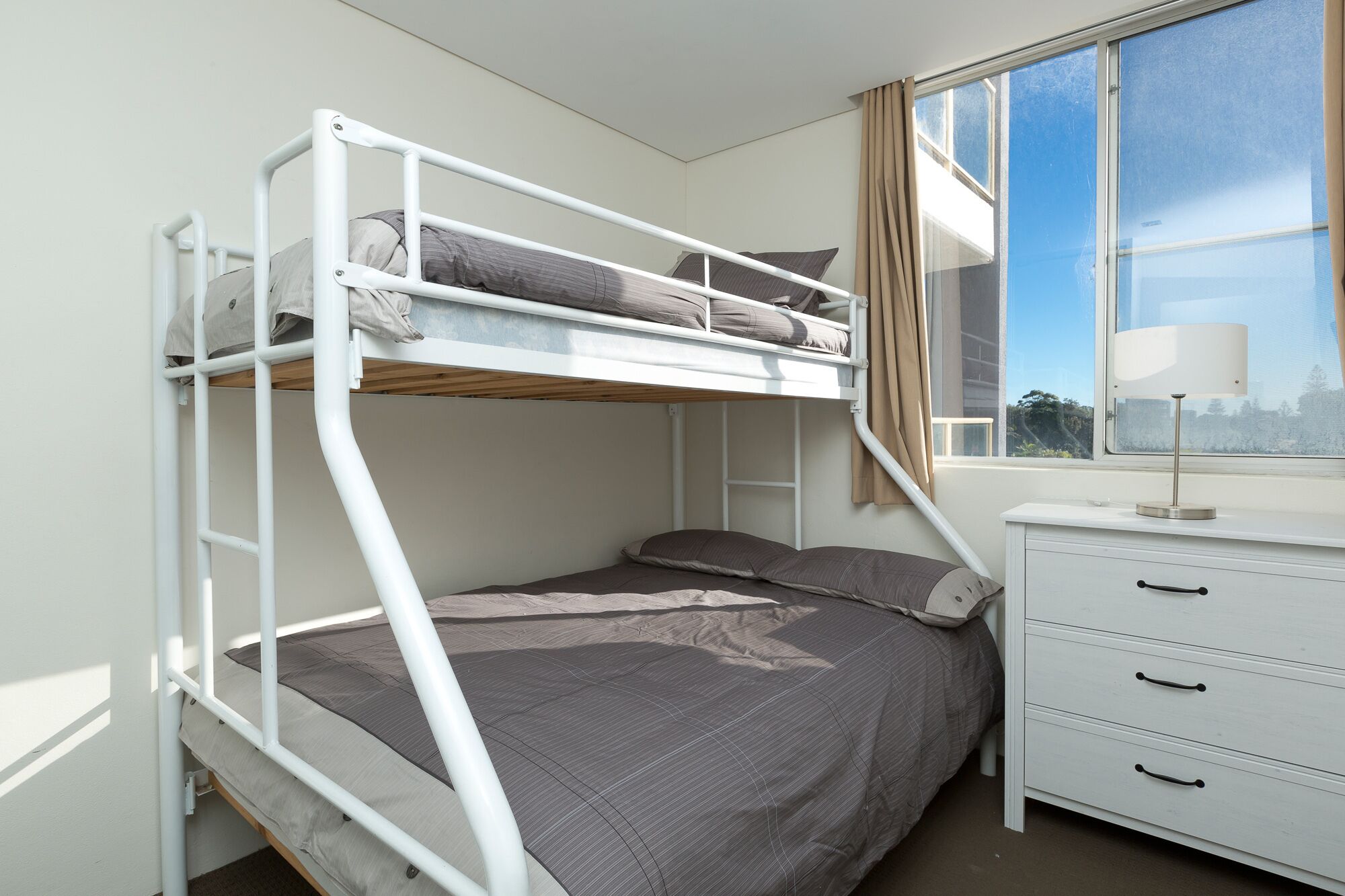 Un lit superposé avec un bureau et des tiroirs, une fenêtre avec des rideaux et une vue sur les arbres.