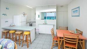 A compact kitchen with a dining area, a refrigerator, a stove, and a view of the sea through a window. - Ebbtide Unit 07 (Forster)