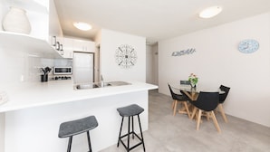 A modern kitchen with white cabinets, a microwave, and a refrigerator. There is a dining area with a glass table and black chairs. - Ebbtide Unit 17 (Forster)