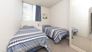 A bedroom with two beds, a window with a curtain, a bedside table with a lamp, and a wall-mounted picture. - Ebbtide Unit 17 (Forster)