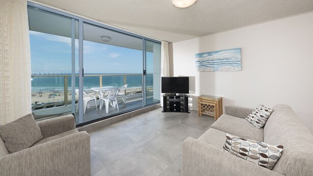 A balcony with a view of the beach and ocean, a flat-screen TV, and a sofa with patterned cushions. - Ebbtide Unit 17 (Forster)