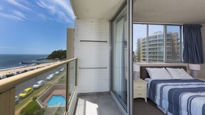 A balcony with a view of the beach and ocean, a bed with a blue and white striped bedspread, and a small window with a curtain. - Ebbtide Unit 17 (Forster)