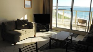 Ett vardagsrum med en soffa, ett soffbord, en tv och ett stort fönster med utsikt över stranden.