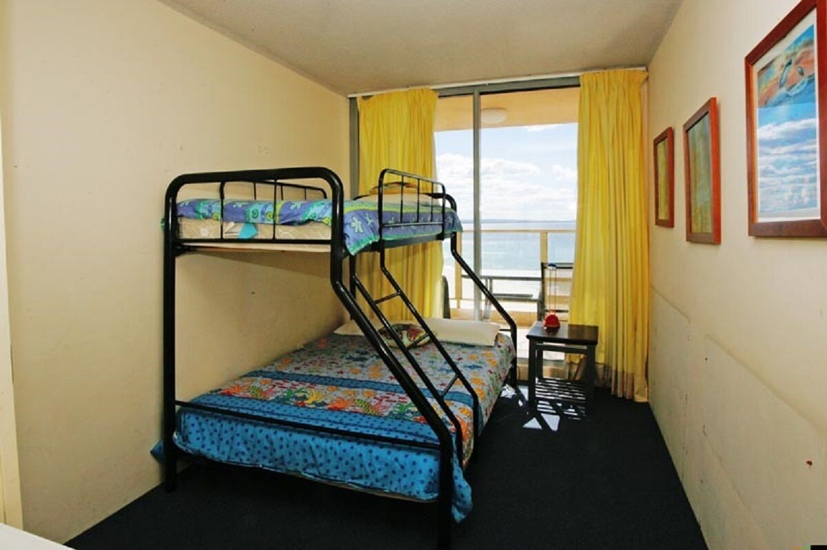 Quarto pequeno com duas beliches, uma mesinha e vista para o mar através de uma janela com cortinas amarelas.