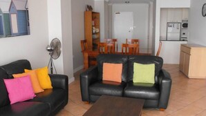 A living room with a black leather sofa, a wooden coffee table, and colorful cushions. There is a dining area with wooden chairs and a table, and a kitchen with white cabinets and appliances. - El Sandi Unit 10 (Forster)