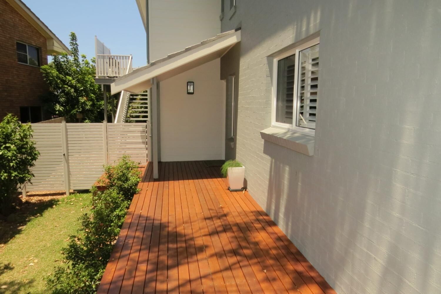 A covered patio area with a wooden deck, a white wall, and a window with shutters.
