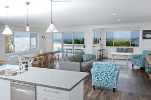 A modern kitchen with a large island, dining area with chairs, and a seating area with a view of the ocean. - Fyne Shores (Forster)