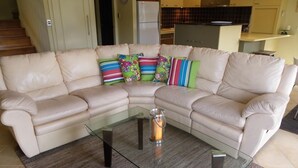 A living room with a beige sectional sofa, colorful pillows, a glass coffee table, and a kitchen in the background. - Gwydir Court 1 (Forster)
