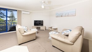A living room with two beige sofas, a flat-screen TV, a ceiling fan, and a painting of birds on the wall. - Heritage 101 (Tuncurry)