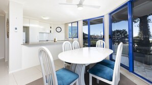 A modern kitchen with white cabinets, a white dining table with blue chairs, and a large sliding glass door leading to an outdoor area. - Heritage 101 (Tuncurry)