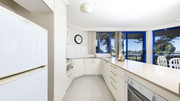 A modern kitchen with white cabinets, a built-in oven, and a large window with blue frames. - Heritage 101 (Tuncurry)