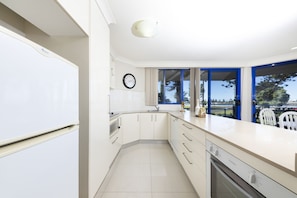 A modern kitchen with white cabinets, a built-in oven, and a large window with blue frames. - Heritage 101 (Tuncurry)