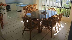 A dining area with a round glass table surrounded by wicker chairs, a kitchen with white cabinets, and a view through a window with blinds. - Hi Surf Unit 2 (Forster)