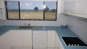A kitchen with a dishwasher, a sink, and a window with a view of the sea and trees. - Hi Surf Unit 4 (Forster)