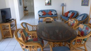 A living room with a round glass table surrounded by wicker chairs, a blue sofa with colorful cushions, a television on a wooden stand, and artwork on the walls. - Hi Surf Unit 4 (Forster)
