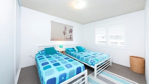 A bedroom with two single beds, a small bedside table, a lamp, a window with blinds, and a wall-mounted picture. - Hibiscus Court Unit 4 (Forster)