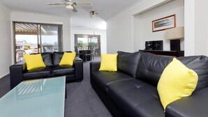 A modern living room with a black leather sofa and glass coffee table, yellow cushions, a ceiling fan, and a view through large windows. - Lake House on Elouera (Forster)