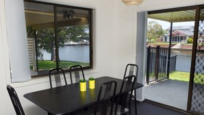 A dining area with a table and chairs, a view of a lake, and a sliding glass door leading to a balcony. - Lake House on Elouera (Forster)