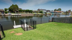 A grassy area with a black metal fence, a small boat dock, and houses in the background. - Lake House on Elouera (Forster)