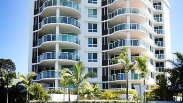 Un hotel de varios pisos con fachada curva, balcones en cada planta y un jardín frontal ajardinado con palmeras.