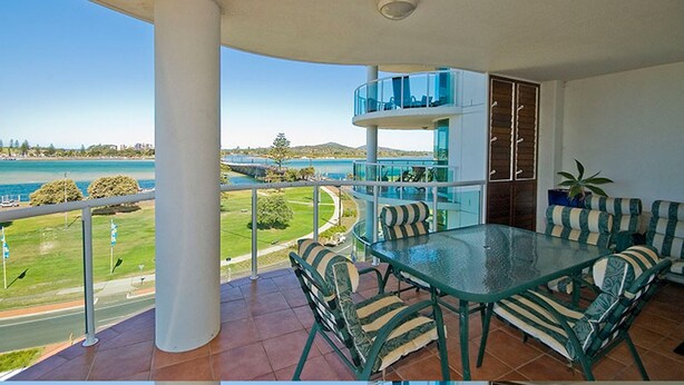 A balcony with a glass table and chairs, overlooking a body of water and greenery. - Mirage Unit 602 (Tuncurry)