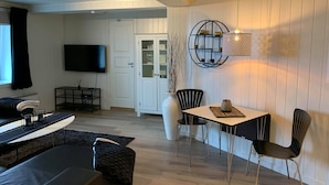 A modern living room with a sofa, a dining table with chairs, a TV mounted on the wall, and a white door. - Notodden Sentrum Apartment No 1 (Notodden)