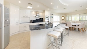A modern kitchen with white cabinets, a black countertop, and stainless steel appliances. There is a dining area with white chairs and a table. - One Mile House (Forster)