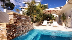 A swimming pool with a stone wall, white lounge chairs, and a palm tree. - One Mile House (Forster)
