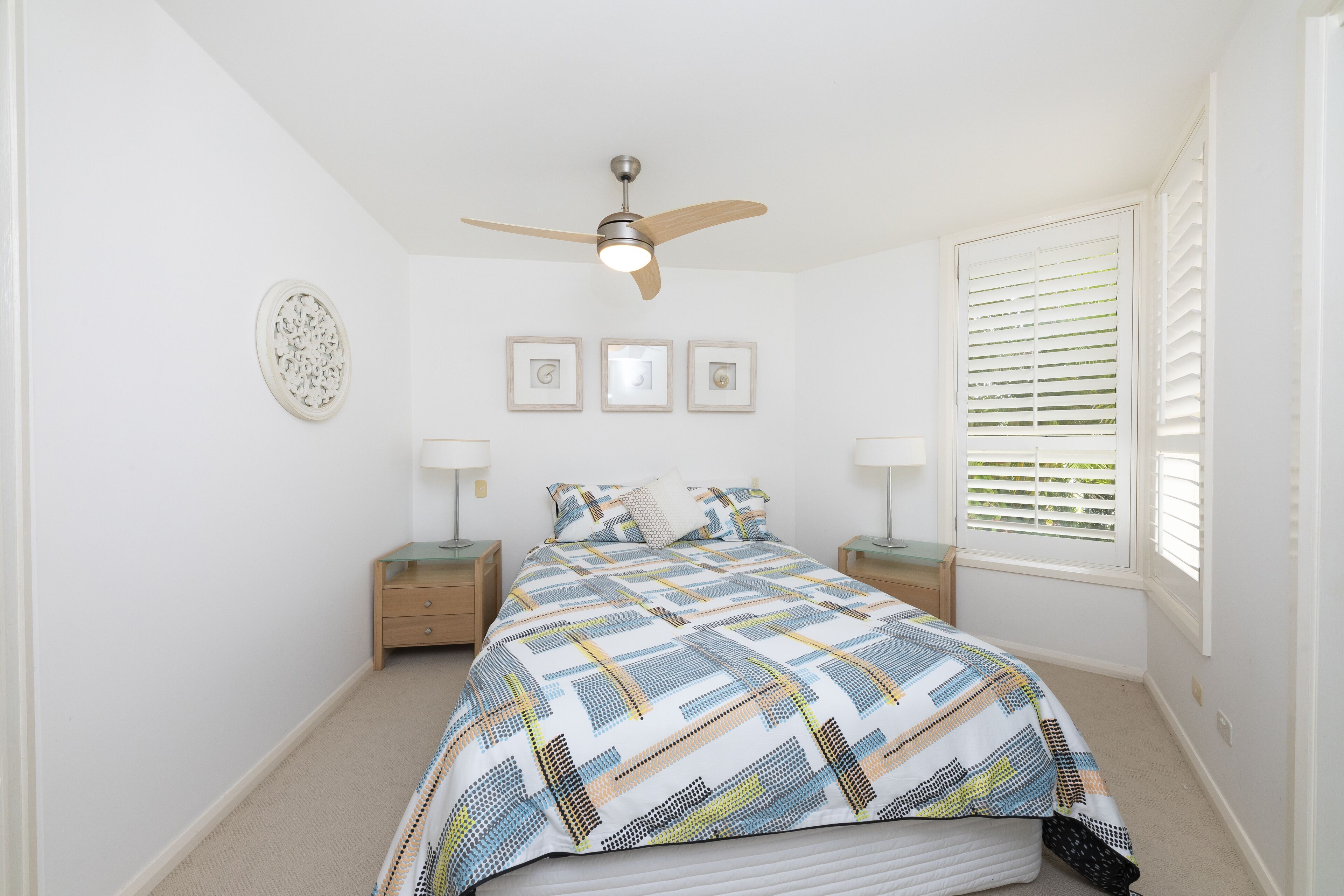 A bedroom with a bed, a ceiling fan, a window with blinds, and a nightstand with a lamp.