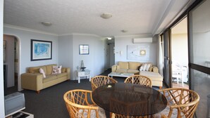 A living room with a round glass table surrounded by wicker chairs, a beige sofa, and a white armchair. There are framed pictures on the walls and a small white side table. - Pinnacle Unit 403 (Forster)