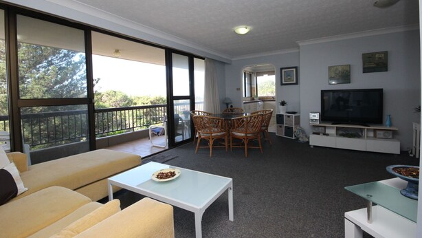 A spacious living area with a balcony, a dining table with chairs, a sofa, a TV, and a small kitchen area. - Pinnacle Unit 403 (Forster)